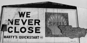 Weathered sign reads We Never Close and Shell, with a damaged wall section on the right.