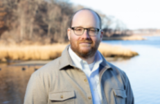 A man in glasses stands by a lake with trees and tall grass in the background.